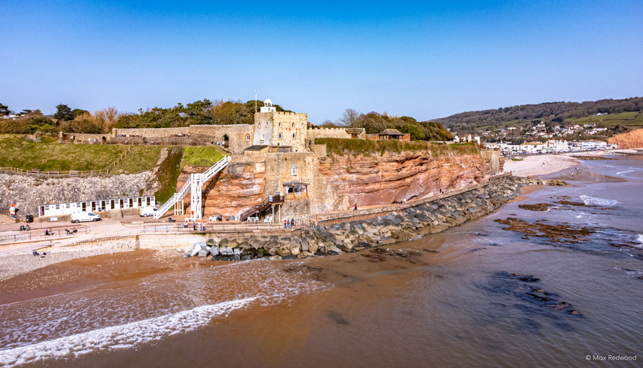 A beach at low tide with an old stone building standing on the shore beneath the red cliffs