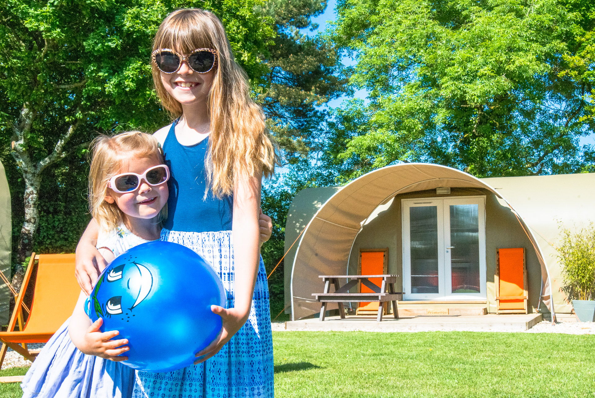 Two children in sunglasses holding a large blue ball on the grass in the sunshine